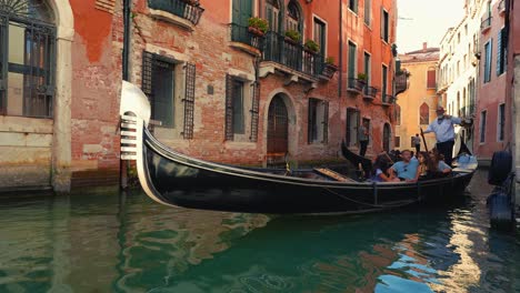 A-gondola-boat,-gondoliere-and-tourists-on-a-canal-in-Venice,-Italy-with-old-houses-and-a-cathedral-church-close-to-San-Marco-and-Rialto-Bridge