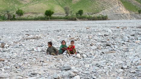 Grupo-De-Tres-Niños-Pequeños-Sentados-En-El-Lecho-Seco-Del-Río-En-Baluchistán,-Pakistán