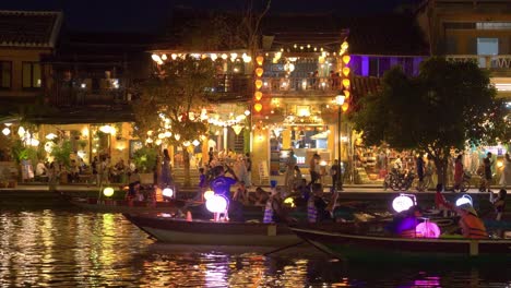 Night-boat-rides-for-tourists-on-Hoai-river-in-ancient-town-Hoi-An,-lanterns-lighting-up-historic-colonial-buildings-fringing-waterfront