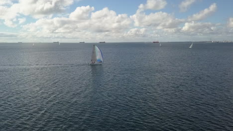 White-sailboat-sails-across-blue-ocean-bay-with-freighters-in-distance