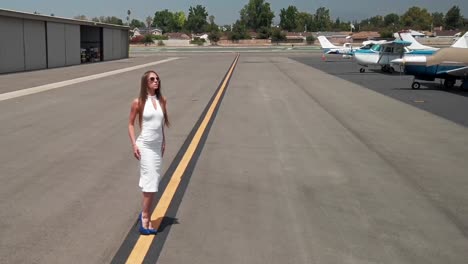 Sexy-Female-Model-in-White-Dress-Posing-to-Camera-in-Front-of-Small-Airplanes-at-Airport,-Slow-Motion