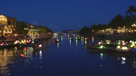 Weite-Nachtaufnahme-über-Dem-Ruhigen-Hoai-Fluss,-Der-Durch-Die-Antike-Stadt-Hoi-An-Schneidet---Traditionelle-Boote-Mit-Bunten-Laternen,-Die-Touristen-Für-Fahrten-Mitnehmen