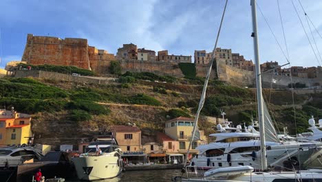Bonifacio-harbor-with-moored-yachts-and-castle-citadel-perched-on-cliff-seen-from-moving-tourist-boat,-Corsica-island-in-France