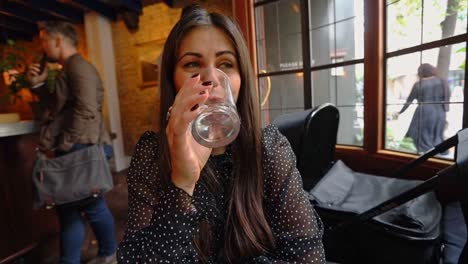 Mujer-De-Pelo-Largo-Bebiendo-Bebidas-En-Un-Moderno-Bar-Londinense-Sentada-Junto-A-La-Ventana-Con-Gente-Pasando-En-El-Fondo