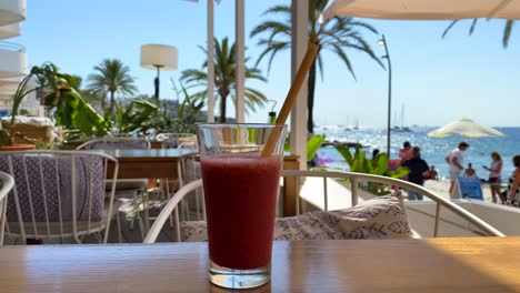 Delicious-red-smoothie-with-fruits-and-berries-by-the-beach-with-palm-trees,-sunny-luxury-holiday-destination,-drink-on-a-tropical-vacation-with-sea-view-in-Ibiza-Spain,-4K-static-shot