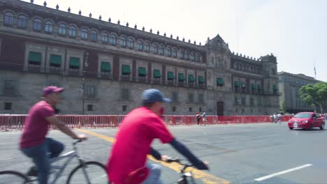Panoramic-view-of-the-government-palace-in-Mexico-City-with-people-walking-and-riding-bicycles-and-cars-driving-by-on-a-sunny-day