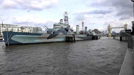HMS-Belfast,-Slow-Motion-of-A-Royal-Navy-Light-Cruiser-Docked-On-River-Thames-On-A-Background-Of-Tower-Bridge-Under-Cloudy-Sky-In-London,-UK