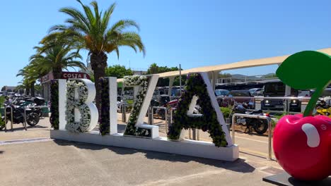 Ibiza-flower-sign-at-the-Ibiza-international-airport-with-big-Pacha-red-cherries-and-palm-trees,-sunny-arrival-in-Spain,-4K-shot