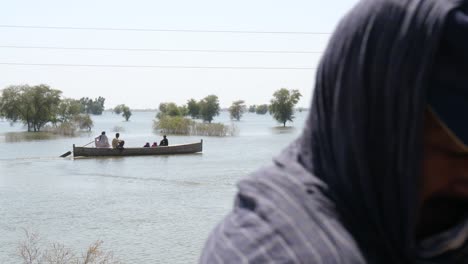Group-Seen-On-Making-Their-Way-Along-Flooded-River-In-Background-With-Blurred-Foreground-Of-Male-Wiping-Face-Due-To-Heat-In-Maher,-Sindh
