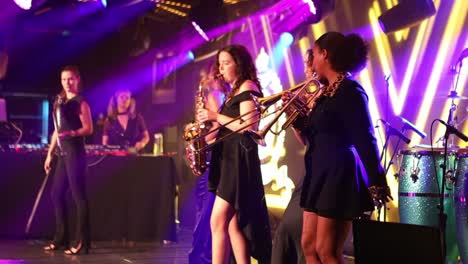 Female-Performers-On-Stage-Singing-And-Playing-Instruments-During-Live-Concert-with-many-colorful-lights-in-the-background-In-London,-UK