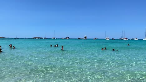Epic-summer-dream-vacation-destination-in-Formentera-island-Ses-Illetes-beach-Ibiza-Spain,-transparent-turquoise-water-and-luxury-yachts-on-a-white-sand-beach,-people-on-holiday,-4K-panning-right