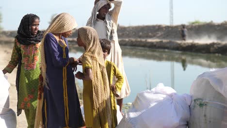 Group-Of-Young-Muslim-Girls-Waiting-Beside-Large-White-Aid-Sacks-Beside-River-In-Maher,-Sindh