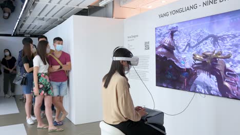 Ein-Kunstbesucher-Verwendet-Ein-Virtual-Reality-Headset,-Um-Mit-Einem-Immersiven-Kunstwerk-Zu-Interagieren,-Während-Andere-Enthusiasten-Und-Besucher-Auf-Der-Digitalen-Kunstmesse-Schlange-Stehen,-Um-Kommende-Trends-Zu-Präsentieren