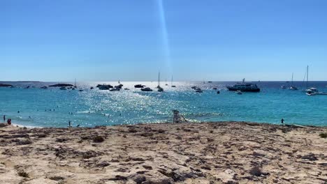 Sunny-summer-dream-vacation-destination-in-Formentera-island-Ses-Illetes-beach-Ibiza-Spain,-transparent-turquoise-water-and-luxury-yachts-on-a-white-sand-rocky-beach,-people-on-holiday,-4K-shot