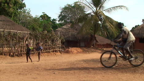 Susana-village-in-northern-Guinea-Bissau,-with-houses-made-of-mud-and-straw,-with-three-children-walking-and-two-adults-passing-by-on-bicycles,-beautiful