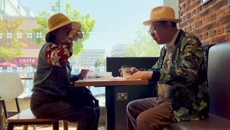 Romantic-elderly-Asian-man-and-woman-wearing-hats-on-vacation,-have-lunch-together-eating-and-drinking-in-a-pub