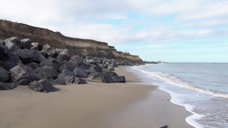 Waves-roll-in-from-the-North-Sea-on-to-a-beach-protected-by-a-sea-defence-of-granite-rocks-on-the-Norfolk-coastline-damaged-by-coastal-erosion