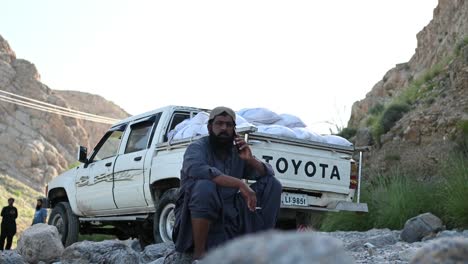 Pakistani-Muslim-Male-Sat-Beside-River-Talking-On-Mobile-Phone-With-Cigarette-In-Hand-In-Sindh-With-Parked-Truck-With-Sacks-Of-Aid-In-Back