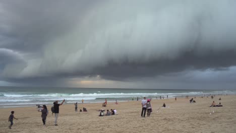 Panning-left-shot-across-coastal-beach-at-surfers-paradise,-thick-layer-of-ominous-dark-storm-clouds-sweeping-across-the-sky,-people-watching-apocalypse-like-weather-at-Gold-Coast,-Queensland