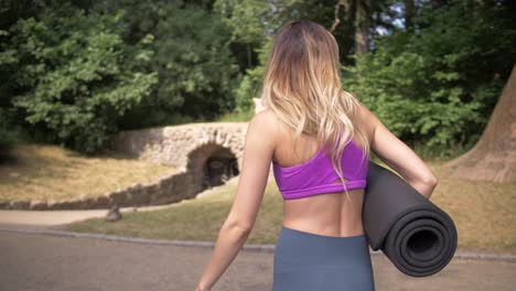 Thin,-fit-blonde-woman-adjusts-her-hair-as-she-walks-toward-a-stone-arch-bridge-in-a-park-carrying-a-yoga-mat