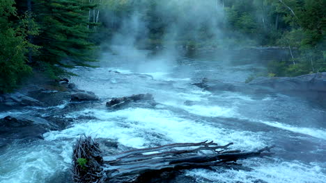Aerial-drone-shot-over-the-dark-misty-forest-stream-at-Big-Wilson-Falls-with-logs-in-the-waterfall-at-dusk-in-Maine