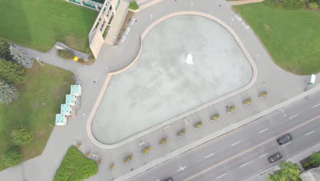 Slowly-moving-downward-footage-from-above-of-a-pond-with-a-fountain-before-a-large-building-with-a-blue-roof