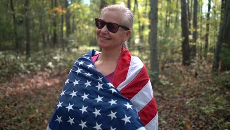 Closeup-of-pretty,-blonde-woman-wrapping-an-American-flag-around-her-and-smiling-in-a-sunlit-forest