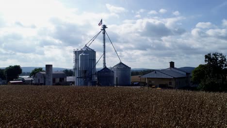 Aufsteigende-Luftbildkamera-Mit-Blick-Auf-Maisfelder-Und-Getreidesilos-Mit-Amerikanischer-Flagge-An-Der-Spitze