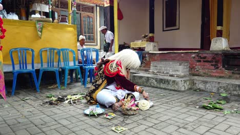 Masked-Character-Performs-Praying-Ceremony-in-Balinese-Temple,-Topeng-Sidakarya-Sitting-on-the-Floor-Manipulating-Sacred-Elements-with-his-Hands,-Water,-Flowers-and-Offerings
