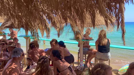 People-sitting-at-a-beautiful-beach-bar-and-enjoying-fun-summer-drinks,-dreamy-holiday-destination-with-sea-view-in-Ibiza-Spain,-4K-shot