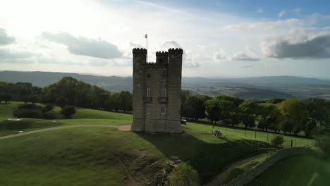 Dramatic-low-aerial-orbit-shot-with-lens-flares-of-Broadway-tower,-an-English-landmark-stood-atop-Beacon-Hill-in-the-Cotswolds,-UK