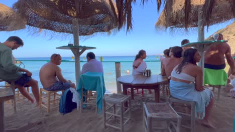 People-sitting-at-a-beautiful-beach-bar-and-enjoying-summer-drinks,-dreamy-holiday-destination-with-sea-view-in-Ibiza-Spain,-friends-and-family-hangout,-4K-shot