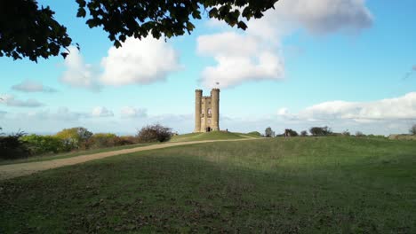 Dramatic-aerial-reveal-shot-of-Broadway-tower,-Cotswolds,-UK,-Reveal-from-tree,-rising-shot-over-turrets-and-union-jack-flag
