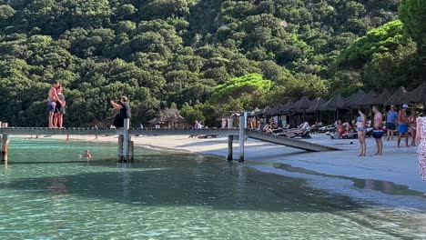 Woman-takes-picture-of-standing-couple-on-wooden-pier-with-phone-at-Santa-Giulia-beach-in-Corsica-island,-France