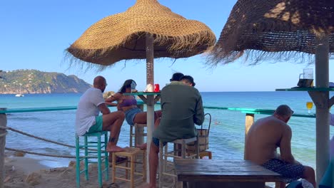 Gente-Sentada-En-Un-Hermoso-Bar-En-La-Playa-Y-Disfrutando-De-Bebidas-De-Verano,-Destino-De-Vacaciones-De-Ensueño-Con-Vista-Al-Mar-En-Ibiza-España,-Reunión-De-Amigos,-Tiro-De-4k