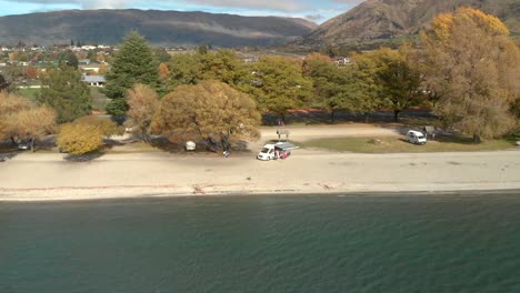 Slowmo-Aerial-Drone-ascending-from-the-beach-overviewing-the-town-and-Lake-Wanaka,-New-Zealand-in-autumn