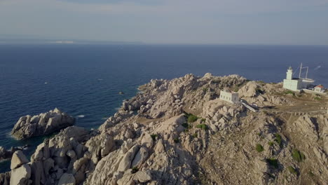 Drone-shot-flying-along-wild-and-rocky-coast-of-north-Sardinia-to-reveal-a-small-white-light-house-with-a-pristine-bay-behind-it