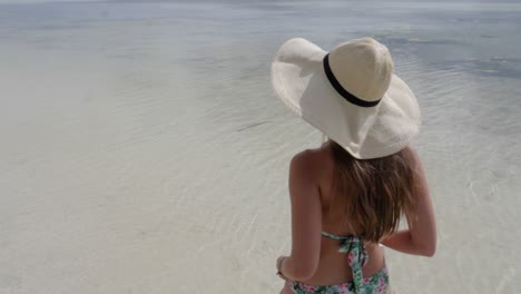 Young-woman-with-bikini-and-hat-walking-away-from-camera-with-book-in-hand-on-beautiful-beach-with-clear-water-in-ultra-slow-motion