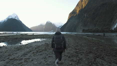 Girl-in-hiking-gear-walking-along-shore-of-Milford-Sound,-New-Zealand