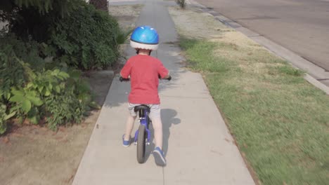 Folgen-Sie-Der-Aufnahme-Eines-Kleinen-Jungen,-Der-Mit-Einem-Blauen-Laufrad-Auf-Einem-Bürgersteig-In-Einer-Wohngegend-Mit-Grünen-Höfen-Fährt