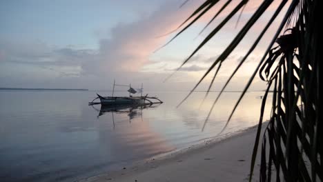 Zeitlupenaufnahme-Eines-Traditionellen-Philippinischen-Bangka-Bootes-Auf-Ruhigem-Wasser-In-Der-Abenddämmerung-Vom-Strand-Hinter-Palmen-Aus-Gesehen