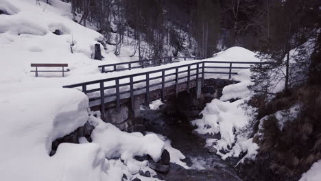 Luftaufnahme-Einer-Kleinen-Brücke-über-Einen-Bach-In-Einem-Verschneiten-Tal-In-Den-Alpen,-Kleinwalsertal,-österreich-Drohne-Hebt-Sich