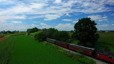 Tren-De-Vapor-Resoplando-A-Lo-Largo-De-Tierras-De-Cultivo-Amish-En-Un-Día-Soleado-De-Verano-Visto-Por-Un-Dron