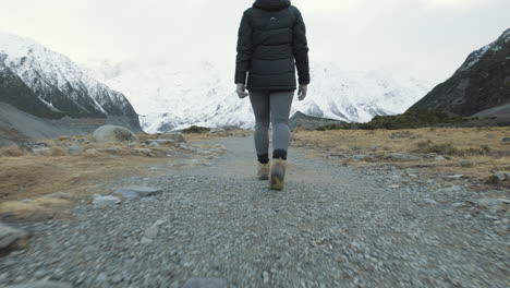 A-low-angle-follow-shot-from-behind-of-a-woman-hiking-between-snow-capped-mountains-on-a-cold-winters-morning-in-New-Zealand