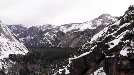 Aerial-Drone-footage,-flyover-cliff-and-drop-off-over-Yosemite-valley-revealing-mountain-range-blanketed-in-snow-in-winter