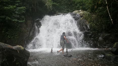 Indonesischer-Mann-Baut-Steinhaufen-In-Einem-Fluss-Vor-Einem-Wasserfall