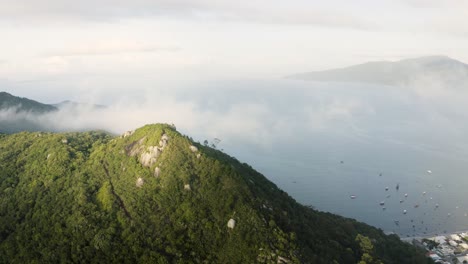 drone-panning-a-cloudy-peak-of-a-tropical-forest-mountain-in-a-summer-day-with-the-ocean-on-the-background