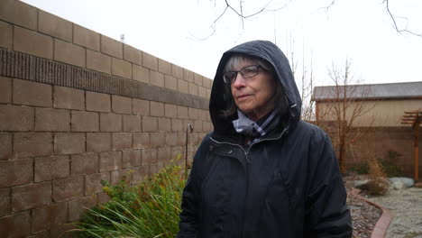 A-beautiful-old-woman-in-glasses-walking-in-a-rain-storm-wearing-a-raincoat-and-hood-as-raindrops-fall-in-slow-motion
