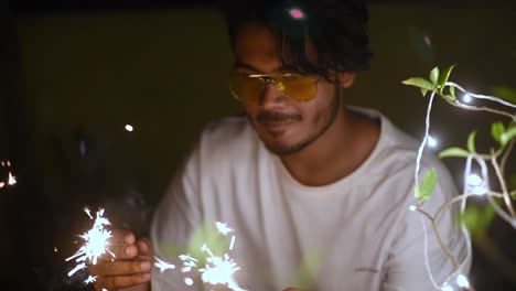 Young-happy-Asian-male-celebrates-Christmas,-Diwali-or-New-Year-party-by-playing-with-fireworks-and-fire-crackers-beside-decorated-lights-on-plant