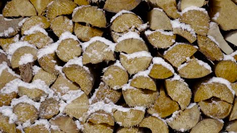 Long-downward-pan-over-cut-logs-of-wood-stacked-up,-covered-in-snow-with-snowflakes-flying-through-the-frame